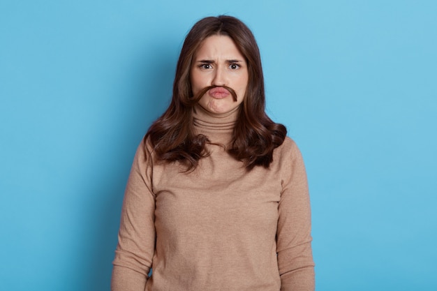 Belle jeune femme avec la moustache de ses cheveux avec un regard sérieux, fronçant les sourcils, vêtue d'un col roulé beige, se dresse contre le mur bleu.