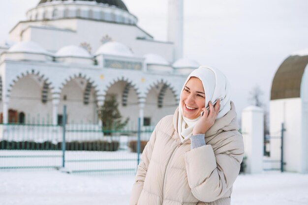 Belle jeune femme musulmane souriante en foulard dans des vêtements légers à l'aide de mobile contre la mosquée