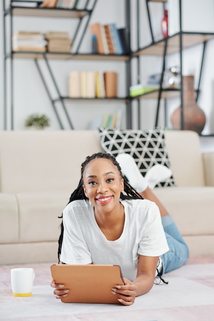 Belle jeune femme noire avec des dreadlocks allongée sur le sol avec une tablette numérique dans les mains et souriant