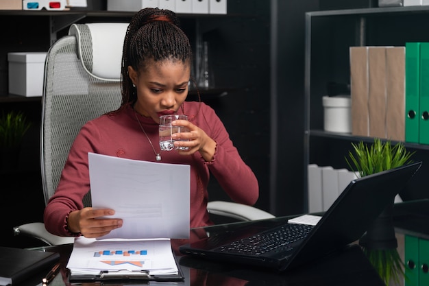 Belle jeune femme noire tenant un verre d'eau et travaillant avec des documents à table au bureau