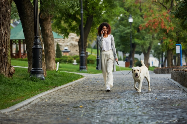 Belle jeune femme occupée marchant avec chien et parler au téléphone