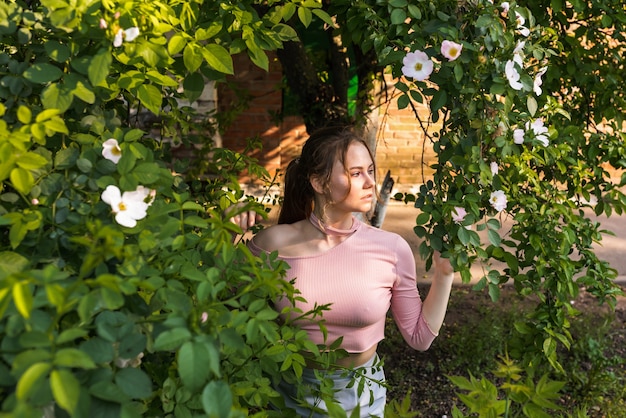 Belle jeune femme à la peau parfaite dans une tenue rose posant près de roses en fleurs dans le jardin