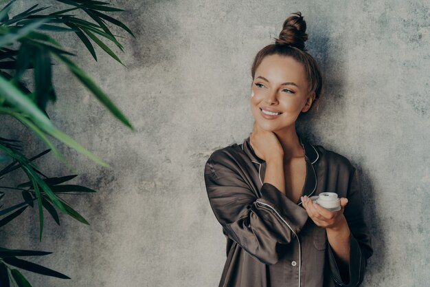 Belle jeune femme avec une peau saine et éclatante souriant largement tout en tenant une crème pour le visage