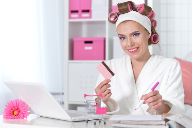 Photo belle jeune femme en peignoir blanc assis à table avec ordinateur portable et faire des achats en ligne