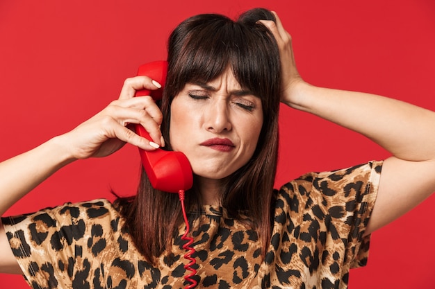 belle jeune femme pensante confuse vêtue d'une chemise imprimée d'animaux posant isolée sur un mur rouge parlant par téléphone.