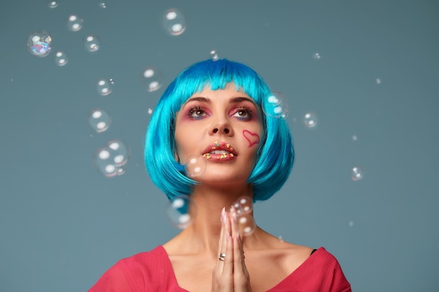 Belle jeune femme avec une perruque bleue et un maquillage lumineux dans des mains de bulles de savon tient au menton fait un vœu Fille de modèle de mode avec un maquillage de couleur créatif Concept de maquillage beauté femme