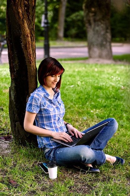 Belle jeune femme pigiste à l'aide d'un ordinateur portable assis sous l'arbre dans le parc.