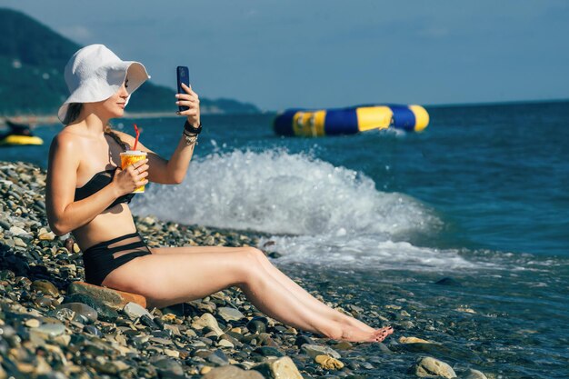 Belle jeune femme sur la plage près de la mer avec un cocktail dans ses mains fait un selfie
