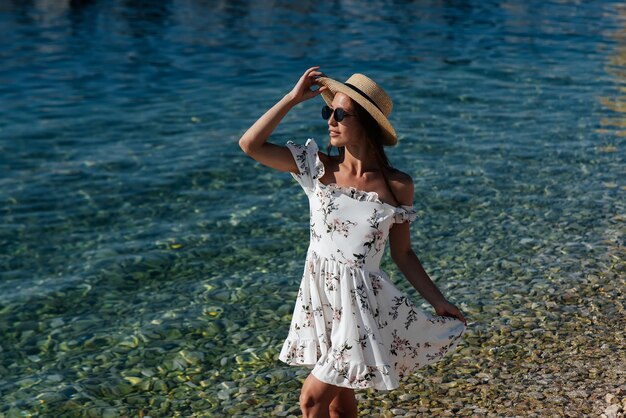 Une belle jeune femme portant un chapeau, des lunettes et une robe légère se promène le long du rivage de l'océan sur fond d'énormes rochers par une journée ensoleillée Tourisme et voyages touristiques