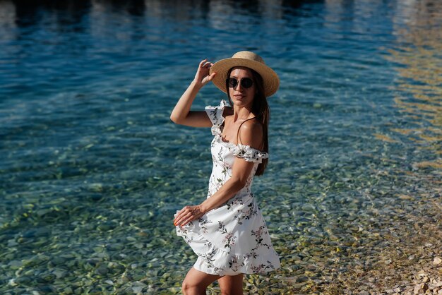 Une belle jeune femme portant un chapeau, des lunettes et une robe légère se promène le long du rivage de l'océan sur fond d'énormes rochers par une journée ensoleillée Tourisme et voyages touristiques