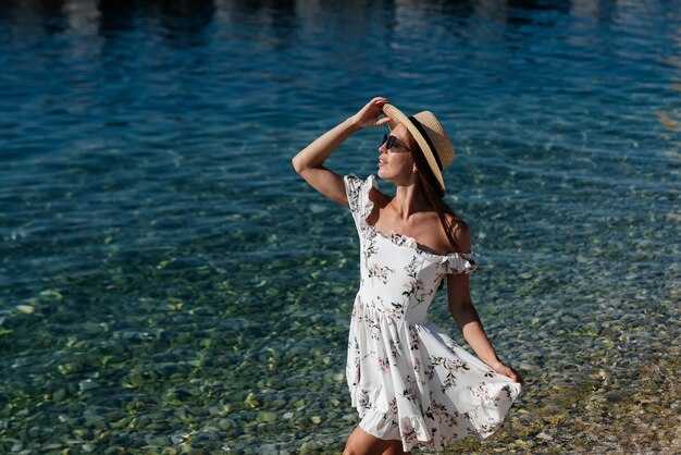 Une belle jeune femme portant un chapeau, des lunettes et une robe légère se promène le long du rivage de l'océan sur fond d'énormes rochers par une journée ensoleillée Tourisme et voyages touristiques