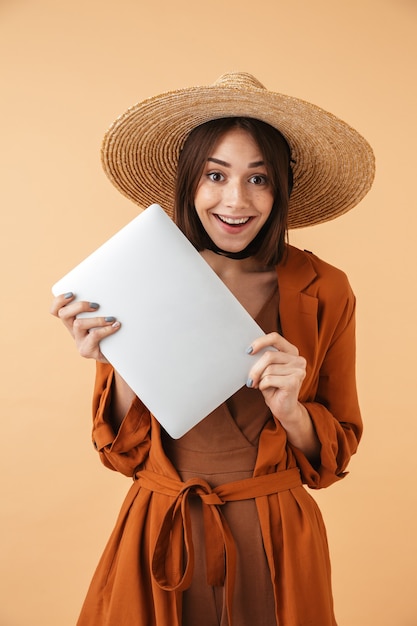 Belle jeune femme portant un chapeau de paille debout isolé sur un mur beige, tenant un ordinateur portable