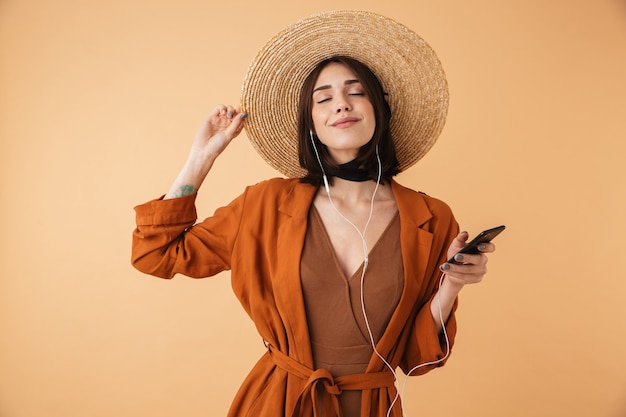 Belle jeune femme portant un chapeau de paille debout isolée sur un mur beige, écoutant de la musique avec des écouteurs, tenant un téléphone portable