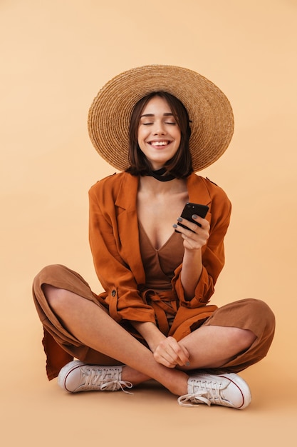 Belle jeune femme portant un chapeau de paille et une tenue d'été assise isolée sur un mur beige, tenant un téléphone portable