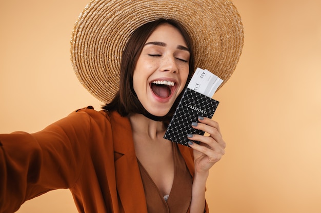 Belle Jeune Femme Portant Un Chapeau De Paille Et Une Tenue D'été Debout Isolée Sur Un Mur Beige, Prenant Un Selfie Tout En Tenant Un Passeport Avec Des Billets D'avion