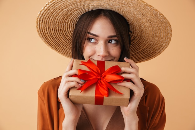 Belle jeune femme portant un chapeau de paille et une tenue d'été isolée sur un mur beige, montrant une boîte-cadeau