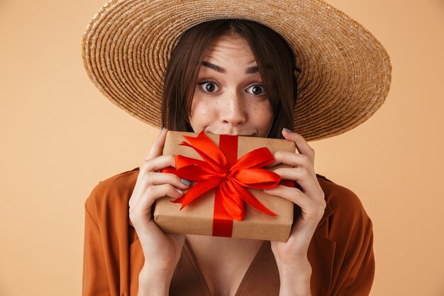 Belle jeune femme portant un chapeau de paille et une tenue d'été isolée sur un mur beige, montrant une boîte-cadeau