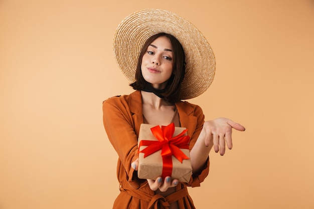 Belle jeune femme portant un chapeau de paille et une tenue d'été isolée sur un mur beige, montrant une boîte-cadeau