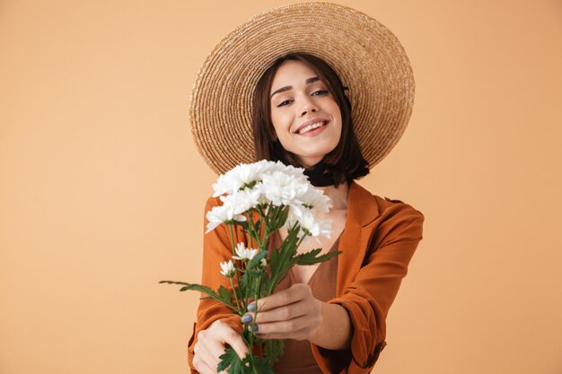 Belle jeune femme portant un chapeau de paille et une tenue d'été isolée sur un mur beige, tenant un bouquet de camomille