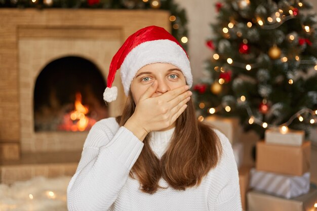 Belle jeune femme portant chapeau de père Noël et pull blanc étant à la maison autour de la décoration de Noël