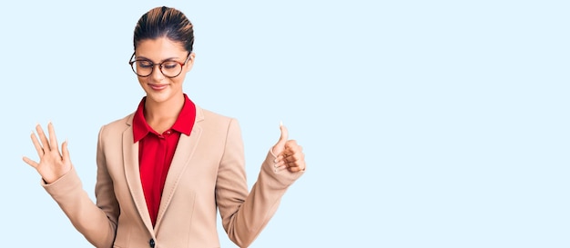 Belle jeune femme portant une chemise d'affaires et des lunettes montrant et pointant vers le haut avec les doigts numéro six tout en souriant confiant et heureux