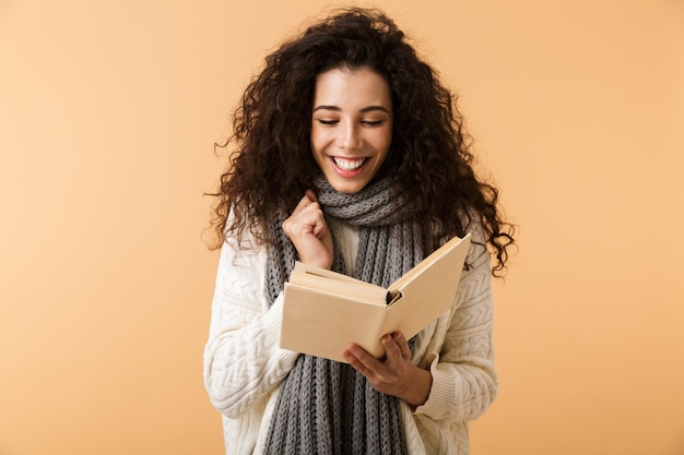 Belle jeune femme portant un foulard d'hiver debout isolé sur un mur beige, lisant un livre