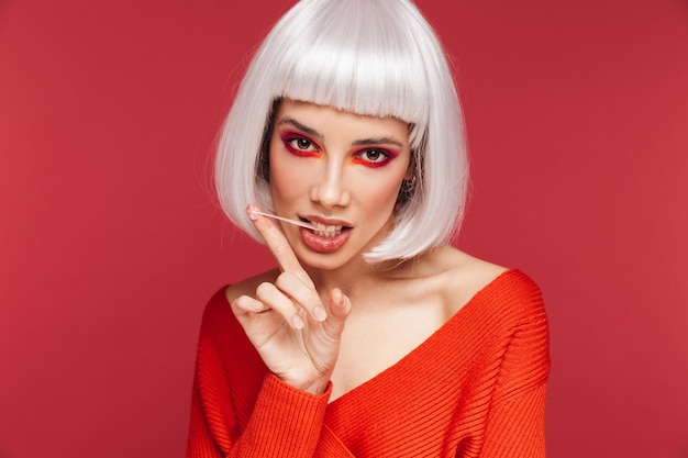 Belle jeune femme portant un maquillage lumineux et une perruque blanche debout isolée sur du rose, mâchant du chewing-gum