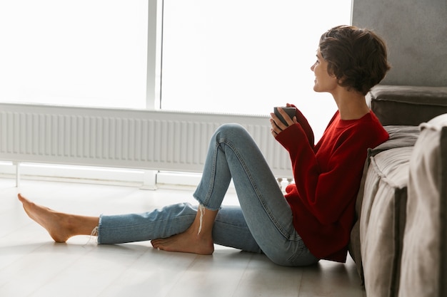 Belle jeune femme portant un pull assis sur un sol à la maison, buvant du thé