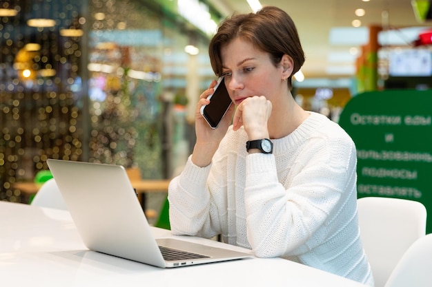 Belle jeune femme portant un sweat-shirt blanc assis dans un centre commercial à une table et travaillant