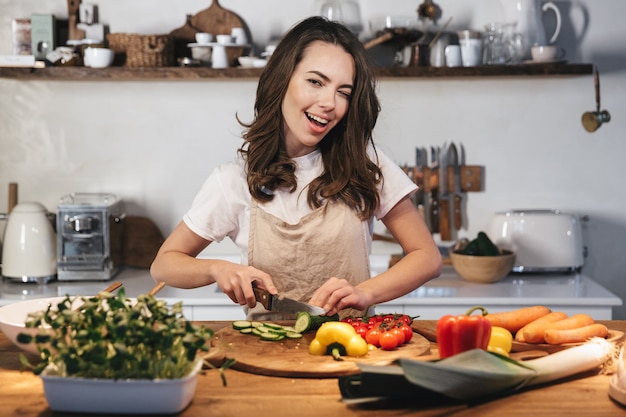 Belle jeune femme portant un tablier cuisinant une salade saine dans la cuisine à la maison, coupant des légumes