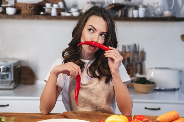 Belle jeune femme portant un tablier cuisinant une salade saine dans la cuisine à la maison, tenant du piment