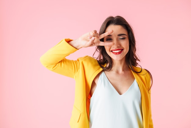 Belle jeune femme portant des vêtements colorés debout isolé sur rose, montrant la paix