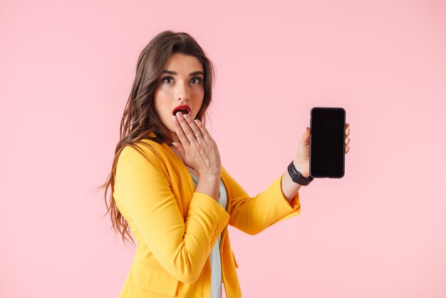 Belle jeune femme portant des vêtements colorés debout isolé sur rose, montrant un téléphone mobile à écran blanc