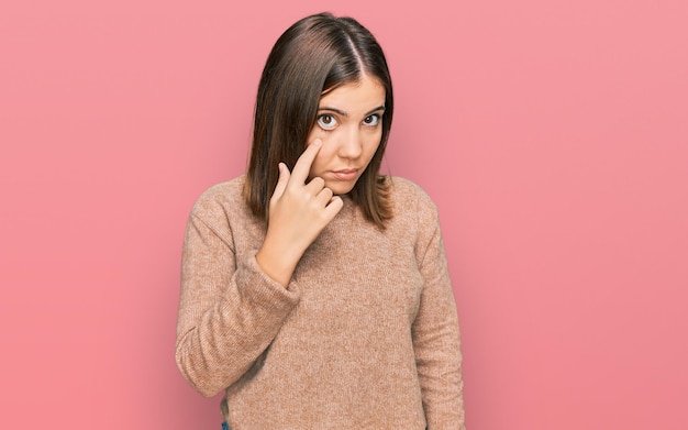 Belle jeune femme portant des vêtements décontractés pointant vers l'œil en vous regardant faire un geste, expression suspecte