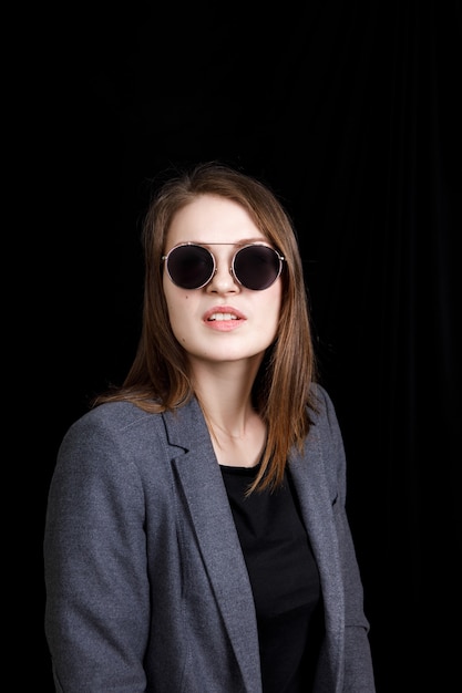 belle jeune femme posant en veste grise et lunettes de soleil sur fond noir