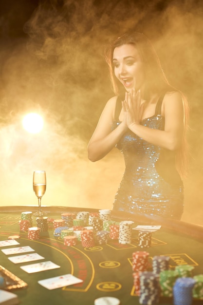 Belle jeune femme pose près d'une table de poker dans un casino de luxe. La passion, les cartes, les jetons, l'alcool, les dés, le jeu, le casino - c'est comme un divertissement féminin. Fond de fumée.