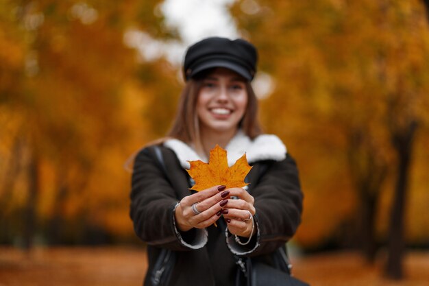 Belle jeune femme positive avec un beau sourire dans un chapeau à la mode avec une visière dans une veste tient une feuille d'érable jaune dans les mains. Jolie fille heureuse montre la feuille d'automne à la caméra et sourit.