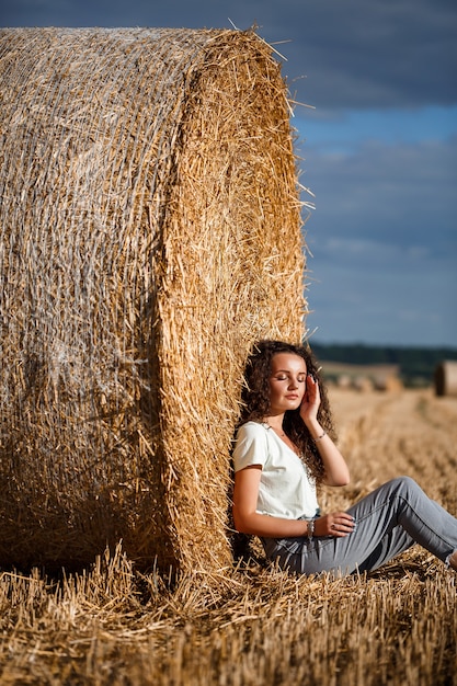 Belle jeune femme près d'une gerbe de foin dans un champ. Vacances dans le village, une fille profitant de la nature dans un champ fauché par une journée ensoleillée