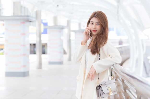 Belle jeune femme professionnelle asiatique en costume blanc sourit et tient un smartphone