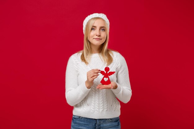 Belle jeune femme en pull blanc, chapeau isolé sur fond rouge, portrait en studio. Mode de vie sain, émotions des gens, concept de saison froide. Maquette de l'espace de copie. Tenez l'ange en papier jouet rouge.