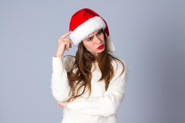 Belle jeune femme en pull blanc avec chapeau de noël rouge se demandant sur fond gris en studio