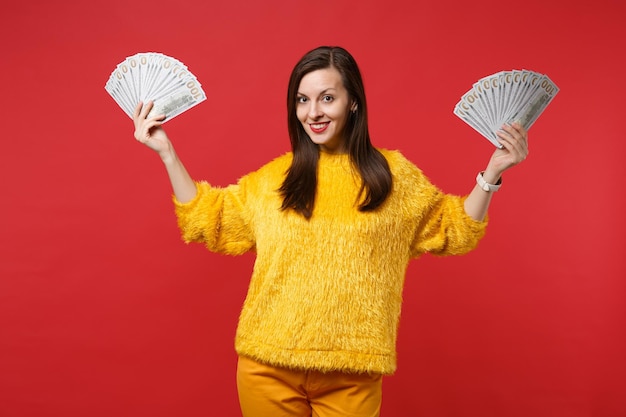 Belle jeune femme en pull de fourrure jaune tenant un ventilateur d'argent en billets de banque en dollars, argent comptant isolé sur fond de mur rouge vif. Les gens émotions sincères, concept de style de vie. Maquette de l'espace de copie.