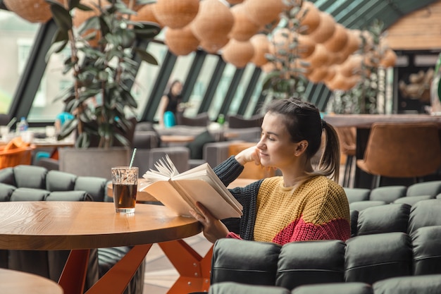 Belle jeune femme en pull orange lisant un livre intéressant au café