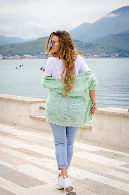 Belle jeune femme en pull oversize, jeans et baskets posant sur le quai de Gaeta en Italie.