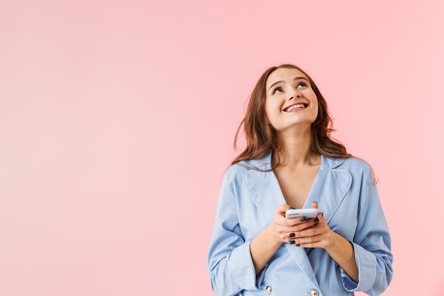 Belle jeune femme en pyjama debout isolé sur fond rose, à l'aide de téléphone mobile