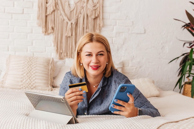 Une belle jeune femme en pyjama est assise sur le lit avec un ordinateur et un téléphone effectue un paiement en ligne Achats en ligne