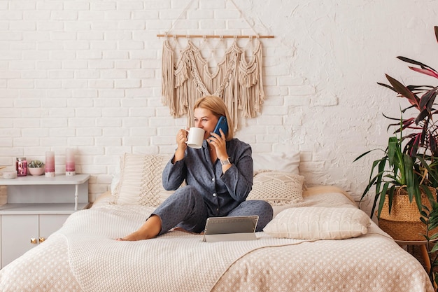 Belle jeune femme en pyjama travaillant sur un ordinateur portable au lit parlant au téléphone Achats en ligne