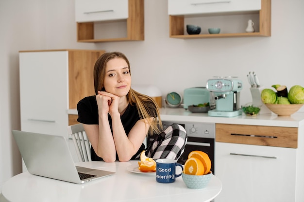 Belle jeune femme en pyjama travaillant avec un ordinateur portable dans la cuisine le matin