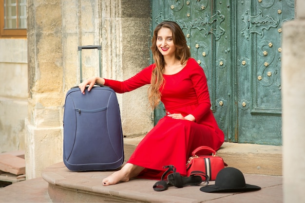 Belle jeune femme de race blanche assise sur les escaliers près de la porte portant un chapeau noir et une longue robe rouge avec valise de voyage et smartphone