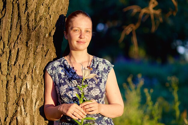 Belle jeune femme de race blanche est debout dans une forêt un jour d'été au coucher du soleil avec une fleur de Lys